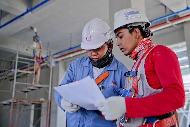 Dos trabajadores de la construcción con cascos y equipo de seguridad revisan los planos en una concurrida obra de Nueva York, destacando las medidas de seguridad para los trabajadores.