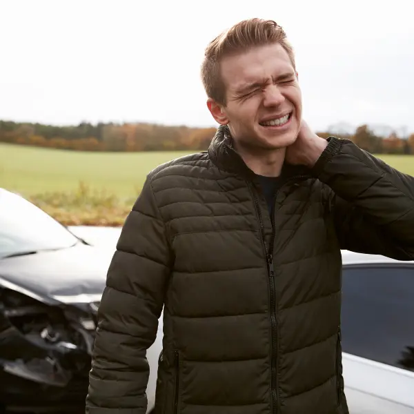 Person lying on the pavement holding their neck in visible pain after a car crash, with a damaged vehicle in the background