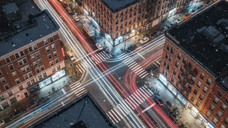 Aerial night view of a New York City intersection with car light trails and crosswalks, representing rising pedestrian accident risks.