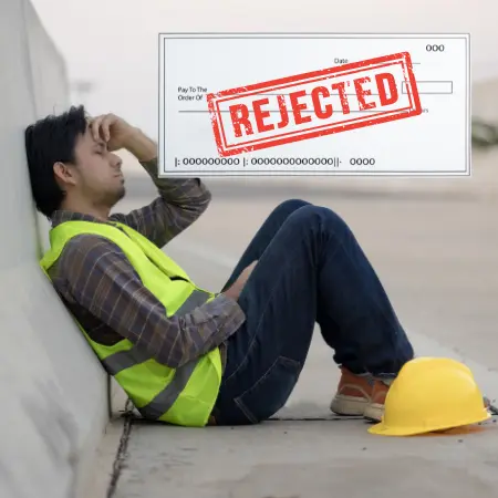 Un obrero latino sentado en el suelo con cara de tristeza, llevándose las manos a la cabeza, mientras en primer plano se ve un cheque rechazado porque la empresa constructora le pilló robando parte de su sueldo.