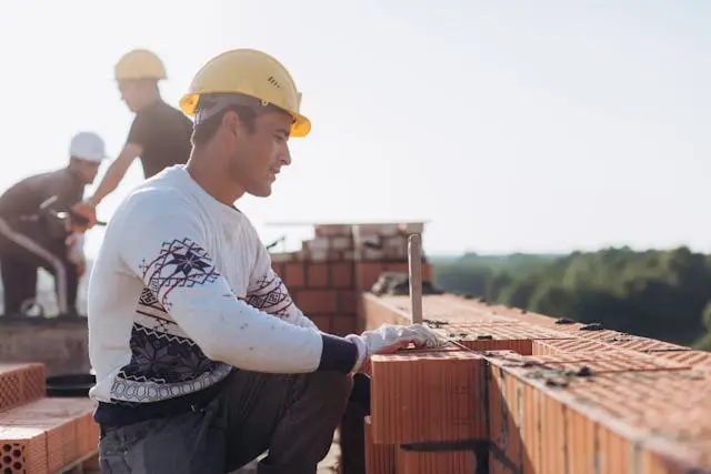 Un trabajador de la construcción de pie sobre un andamio bajo el sol de julio, equipado con material de seguridad, con un cielo azul difuso y un fondo industrial.