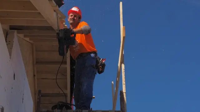 Obrero de la construcción de pie sobre un andamio bajo el sol de julio, equipado con material de seguridad, con un cielo azul difuso y un fondo industrial. Llama la atención sobre las infracciones en las obras de julio, como la falta de equipos de protección contra caídas.