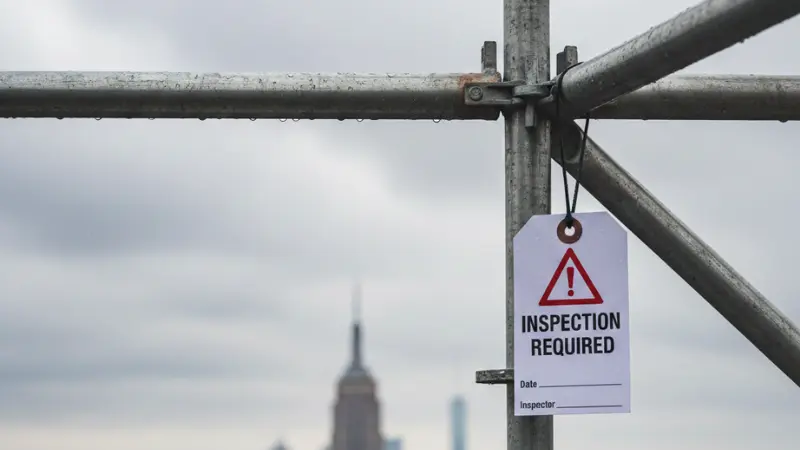 Close-up of metal scaffolding structure in New York City with safety inspection tag under cloudy sky, symbolizing construction and pedestrian safety.