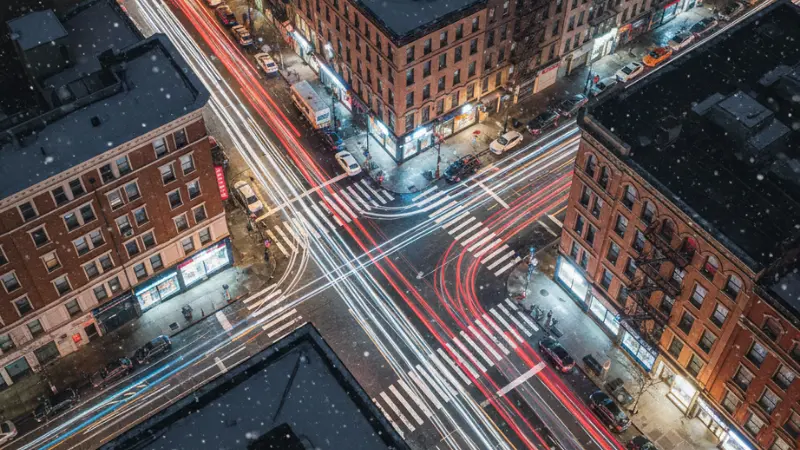 Vista aérea nocturna de una intersección de la ciudad de Nueva York con rastros de luces de automóviles y pasos de peatones, lo que representa el aumento del riesgo de accidentes de peatones.