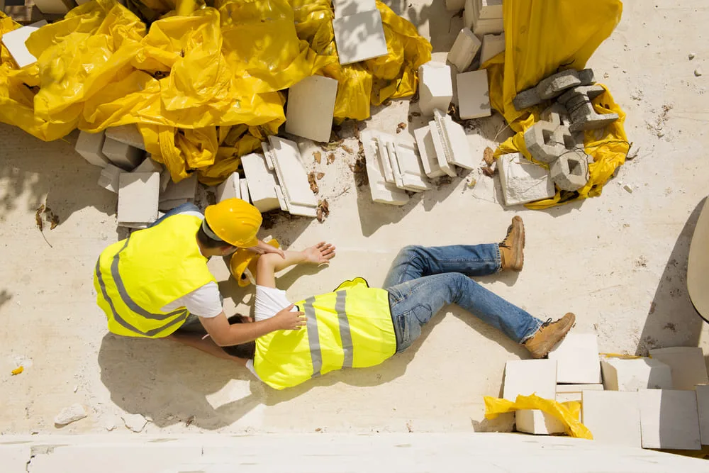njured construction worker lying on the ground while another worker helps him amid tiles and cement blocks at a New York City job site