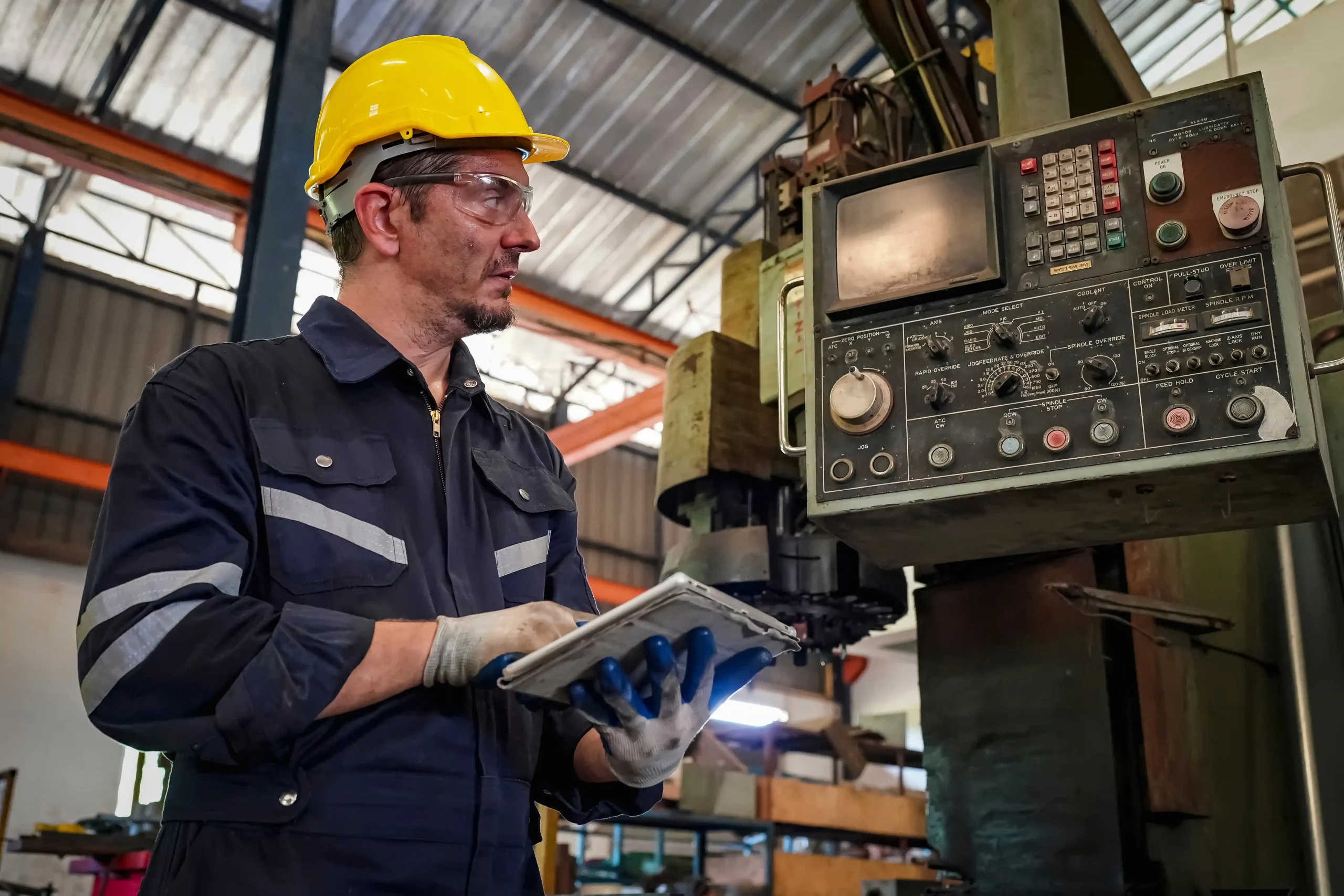 Worker in yellow helmet and safety goggles controlling a machine panel with a tablet in a large industrial workshop in New York.