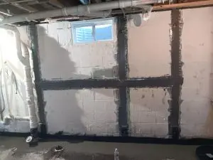 Basement interior with exposed pipes and window, features a cinder block wall reinforced with horizontal and vertical black beams; repair work visible.