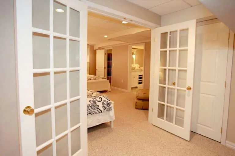 Finished basement interior with double glass doors leading to a cozy guest suite, featuring beds, soft lighting, and a welcoming atmosphere, showcasing basement remodeling options by Basement Repair Specialists.