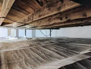 Wooden beams under a house with a vapor barrier covering the ground. Exposed cables and support columns are visible, creating a rustic, utilitarian tone.