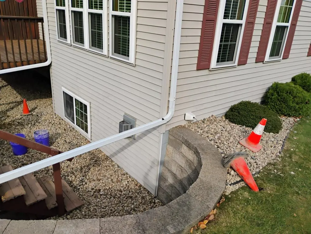 Why Spring Is the Best Time to Waterproof Your Basement Exterior view of a home showcasing a basement window, drainage system, and waterproofing measures, with landscaping elements including gravel and traffic cones, highlighting the importance of basement waterproofing in spring.