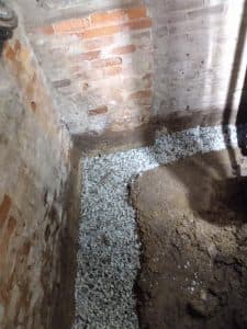Interior basement corner showing gravel drainage system for waterproofing, exposed brick walls, and soil area, illustrating basement repair techniques and moisture prevention.