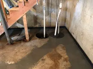 Basement floor with freshly poured concrete, sump pump pipes installed, and wooden stairs in the background, illustrating professional waterproofing solutions for water damage prevention.
