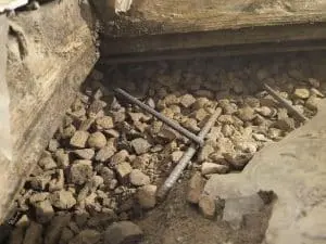 Exposed gravel, soil, and metal reinforcing bars beneath wooden structural beams during basement repair work.