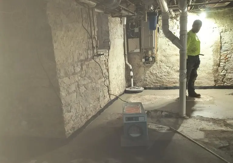 Basement with installed Aprilaire dehumidifier, technician in safety gear, moisture control setup, stone walls, and plumbing visible, illustrating dehumidification services for improved air quality.