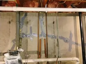Concrete wall with exposed pipes features patched areas sealed with dark blue material. PVC pipes and insulation visible, suggesting repair work in a basement.