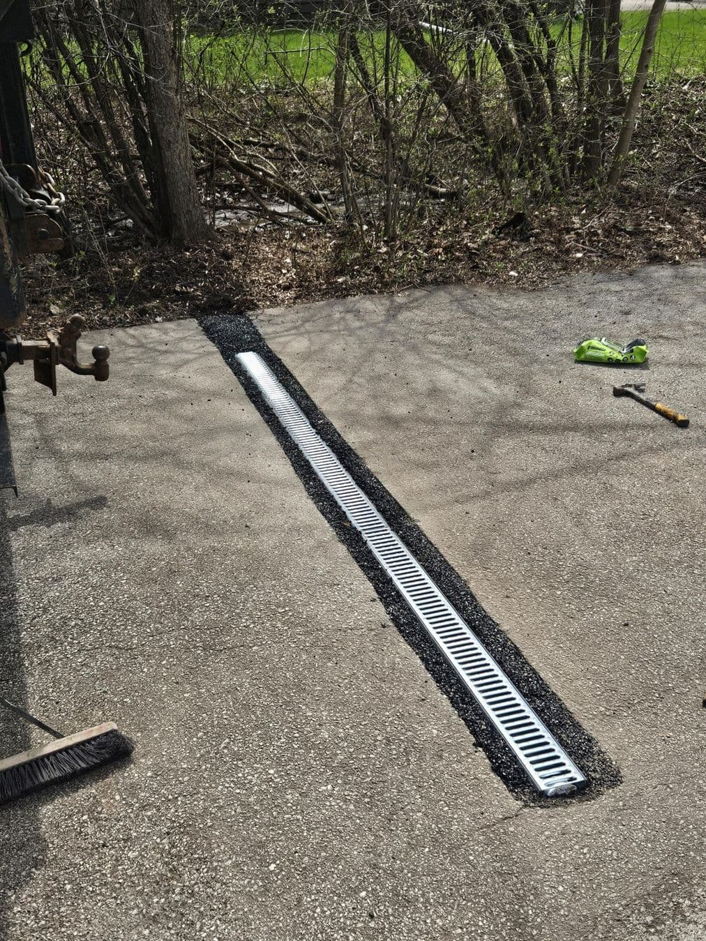 Exterior drain tile system installation on a gravel surface, featuring a long metal grate for efficient water diversion, with tools nearby and surrounding greenery visible.