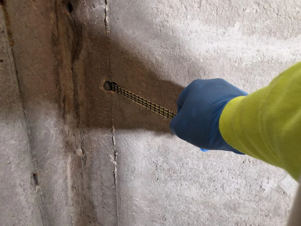 Hand in blue glove using a tool on a crack in a concrete basement wall, illustrating basement repair techniques for waterproofing and structural integrity.