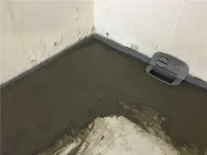 Concrete floor with a newly applied layer in a basement corner, featuring a drainage system component, illustrating interior waterproofing methods to prevent water damage.