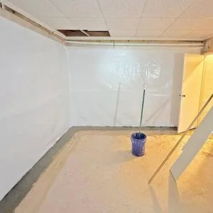Basement interior after waterproofing, featuring white plastic sheeting on walls, clean floor, and a bucket, illustrating effective moisture control.