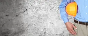 Construction professional in a blue shirt holding a yellow hard hat against a textured gray wall, symbolizing basement foundation repair expertise.