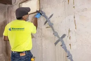Professional applying crack repair techniques on a basement wall, wearing a bright yellow shirt with "Basement Repair Specialists" branding, emphasizing foundation maintenance and repair.
