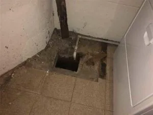Basement floor with visible water damage, exposed plumbing, and a drainage hole, illustrating issues that professional waterproofing services can address.