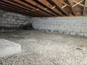 A dimly lit crawl space with exposed wooden beams and concrete block walls. The gravel floor adds a rough texture, emphasizing an unfinished look.