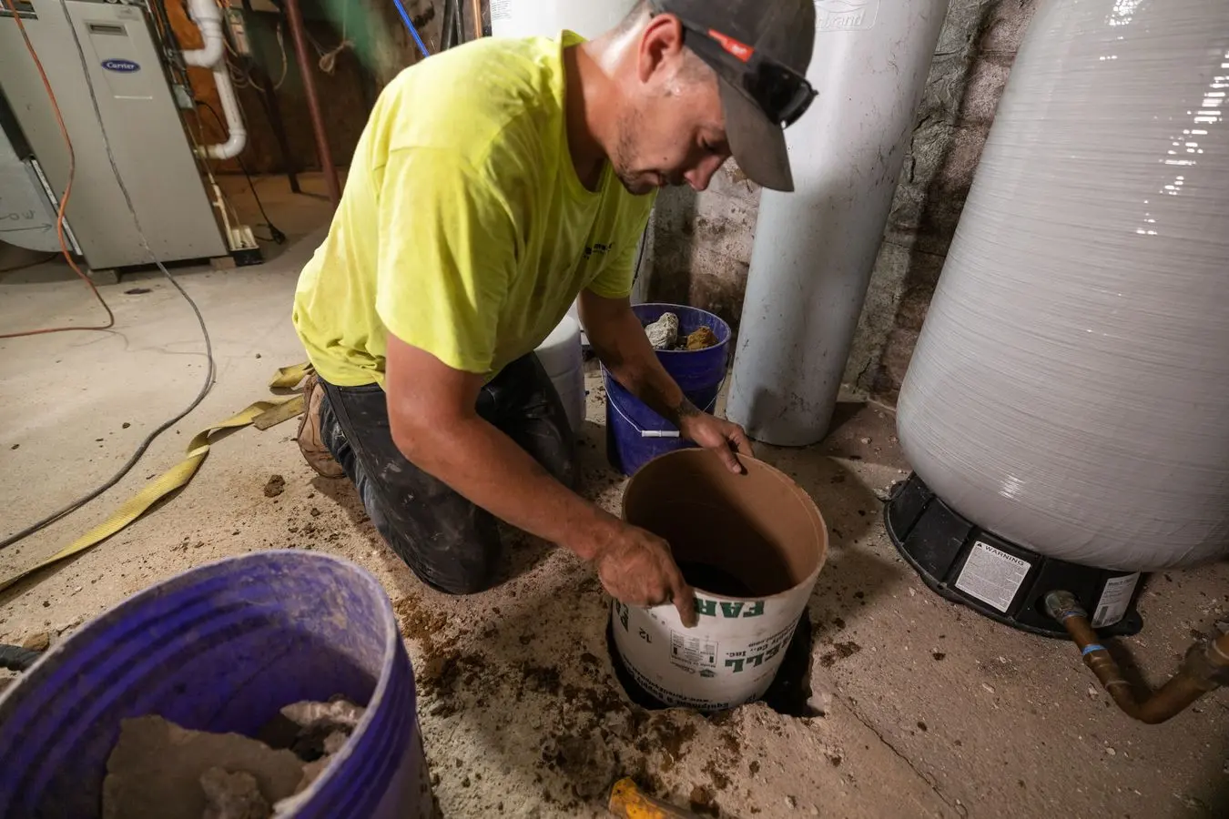 Worker installing a water control system in a basement, handling a drainage bucket, surrounded by tools and equipment for basement repair and waterproofing.
