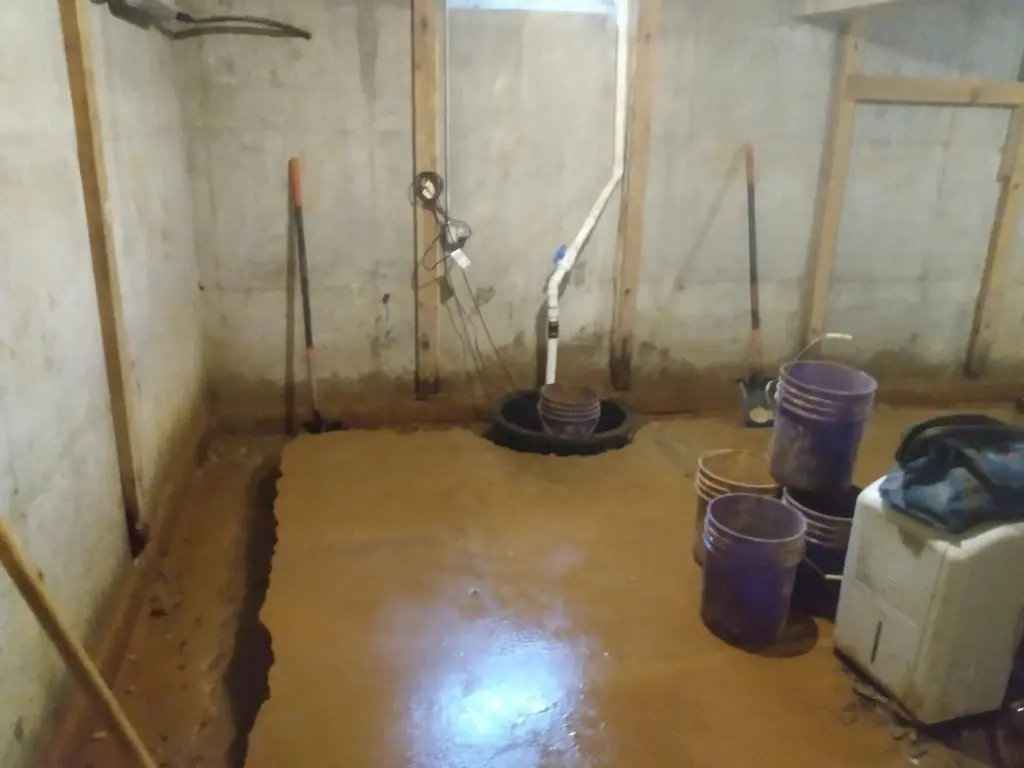 Interior basement scene with wet concrete floor, sump pump installation, and tools for basement waterproofing and repair, highlighting the process of addressing basement leaks.