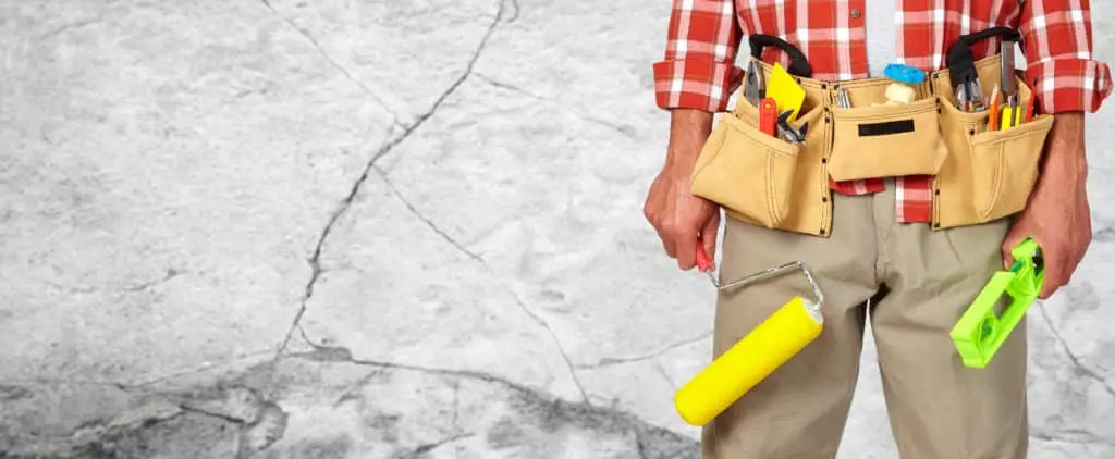 Person wearing a tool belt holding a paint roller and level, standing in front of a cracked concrete wall, representing basement repair and maintenance.