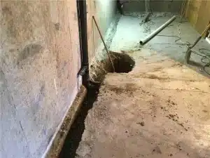 Basement floor showing excavation for waterproofing, with exposed dirt, drainage pipe, and tools, illustrating the process of preventing water intrusion and foundation repair.