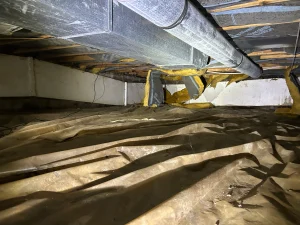 A dimly lit crawl space with exposed ducts and insulation overhead. The floor is covered with a wrinkled, dusty tarp, creating an abandoned, cluttered feel.