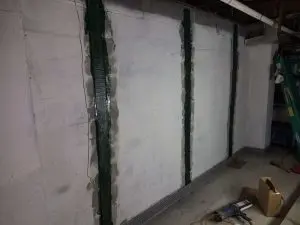 Basement wall with dark vertical reinforcements and light concrete, tools on the floor, a ladder, and exposed pipes above, conveying a work-in-progress.