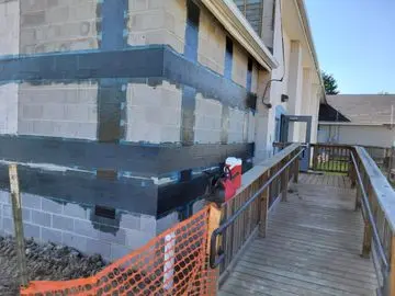 Exterior wall with carbon fiber reinforcement applied for foundation repair, featuring gray cinder block, blue tape, and wooden ramp leading to a door.