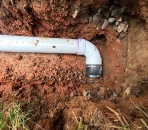 White PVC drainage pipe and elbow fitting installed in an excavated trench surrounded by red clay soil.