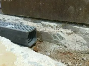 Close-up of a construction site showing a black plastic drainage channel placed in a shallow trench with exposed dirt and concrete. Natural lighting.