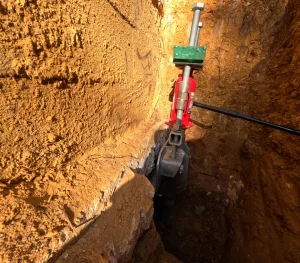 A hydraulic jack is tightly secured between two hard clay walls in a trench. Sunlight casts shadows, highlighting the rough, textured surfaces.