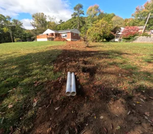 Two white pipes protrude from the ground in a grassy backyard, leading toward a brick house. The area around the pipes is disturbed soil, under a partly cloudy sky.