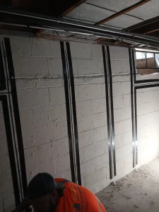 Basement wall with vertical and horizontal black support straps, a small window, and exposed pipes. A person in an orange shirt is partially visible.