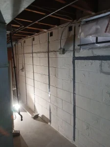 Dimly lit basement with exposed ceiling beams, concrete walls with vertical reinforcement strips, and a bright utility light on the floor.