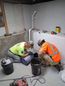 Technicians installing a sump pump system in a basement, focused on equipment and materials, with tools and buckets visible, showcasing foundation repair work by Toledo Basement Repair.