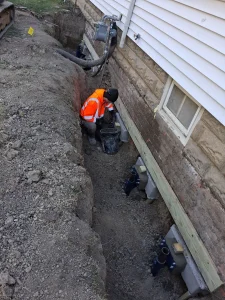 Worker installing a GrateProducts™ Closed Sump System in excavated trench next to a home's foundation, addressing waterproofing and structural issues.