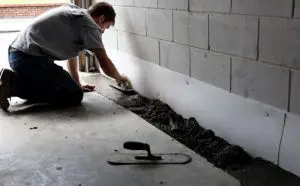 A person kneels on a concrete floor, smoothing wet cement with a trowel against a white wall, suggesting construction or repair work in progress.