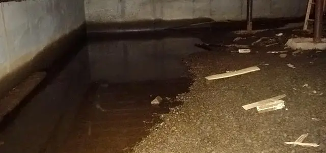 Flooded basement with murky water, debris on gravel floor, and exposed pipes along the ceiling. The scene conveys a sense of neglect and damage.