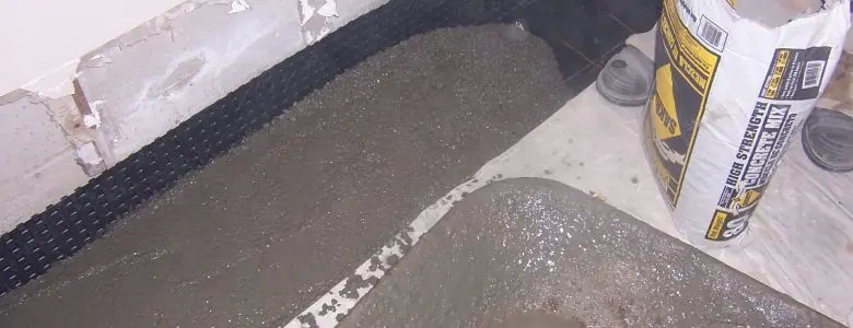 Concrete is being poured along a basement wall with exposed insulation, next to a bag of cement mix. The setting feels industrial and unfinished.
