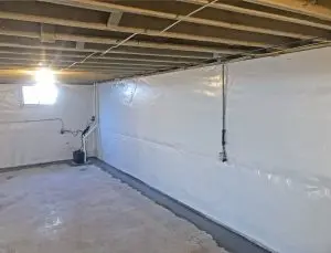 A clean, unfinished basement with exposed wooden ceiling beams and a bright bulb hanging, illuminating plastic-covered walls. A small window and pipes are visible.