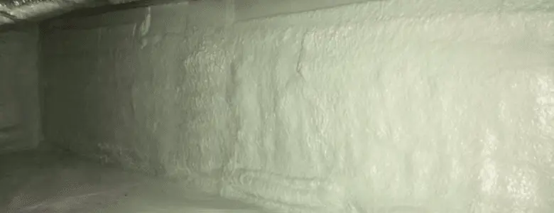 Narrow crawl space with light green spray foam insulation on walls and ceiling, wooden beams, and a clean white plastic sheet covering the floor.