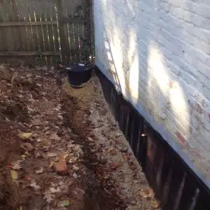 A narrow trench beside a white brick wall is filled with gravel, surrounded by fallen leaves. A dark cylindrical container sits at the trench's end.