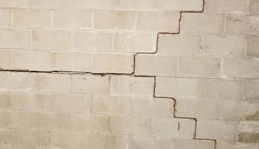 A cracked cinder block wall with a prominent jagged horizontal and step-like vertical crack. The image conveys a sense of structural damage.