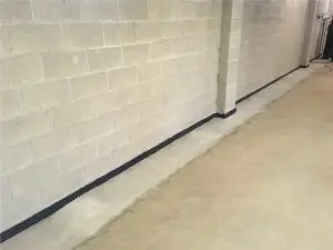 A beige concrete block basement wall with a black drainage strip along the base. The floor is a light tan concrete, creating a clean, minimal appearance.